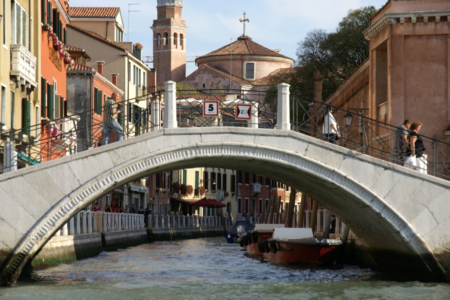 A white stone bridge crosses the entrance to a waterway with three- and four-story buildings on both sides separated from the water by narrow walkways with mooring posts for boats along each edge