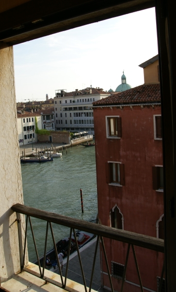 Looking through an open window at a four-story red stucco building and wide waterway with boats moored at posts on either side and buildings on the other side of the canal