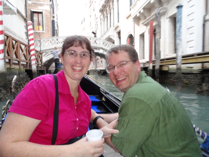 James and Julie smile for the camera as their Gondola approaches a low bridge over a narrow canal with building walls on both sides