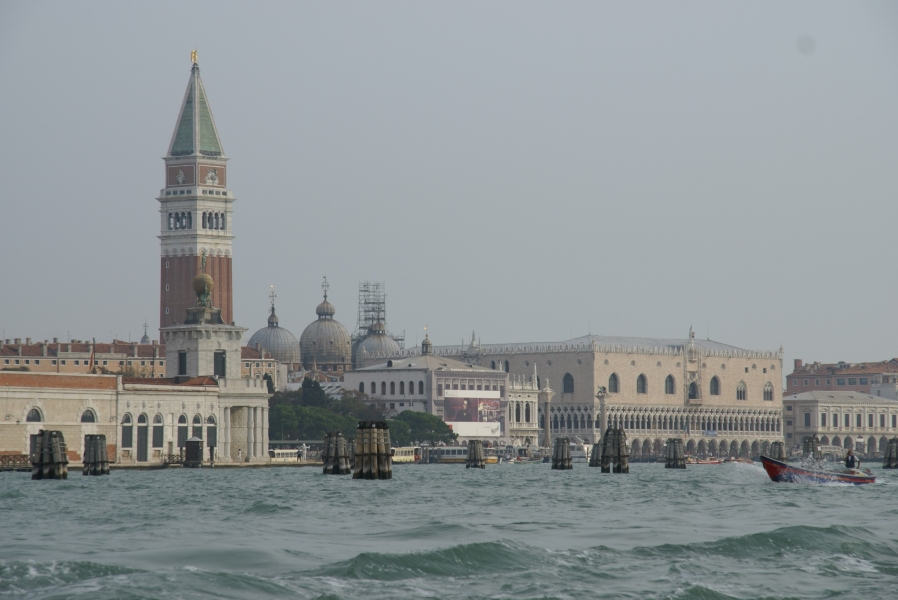 Choppy water moves past mooring piles in front of buildings of various styles near the water including one with ornate white stone arches supporting patterned pink and white blocks and a large square tower of red brick and green roof tiles sticks more than twice as high above the tallest four-story buildings around it