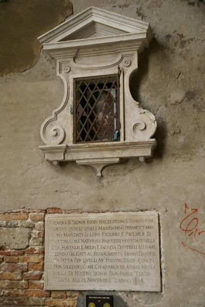 A plaque dated 1548 is embedded in a brick wall below a small, framed opening with a religious marker behind a metal grate