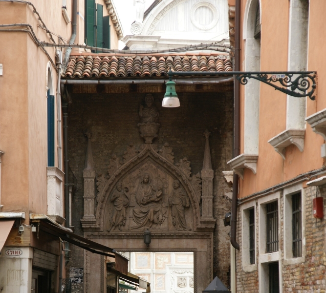 A small terra cotta roof protects stone carvings on a brick wall identifying a historic gate in one of the streets of Venice