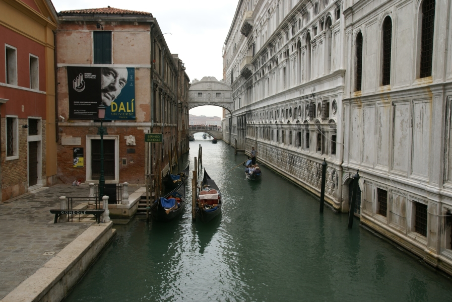 A small white enclosed bridge with carved stone grills in the small windows spans a canal between the second floors of two buildings directly against a canal
