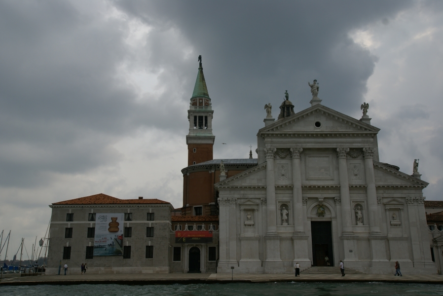 A white stone façade with statues and a single door stands on a small open plaza which ends at the water’s edge stands before a red brick building with a bell tower