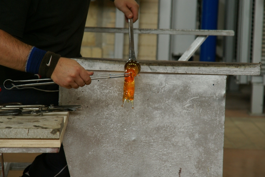 A man uses tools to pull tendrils of glass from a heated blob attached to blow rod