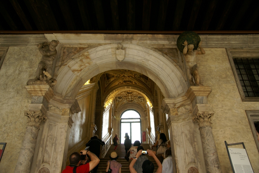 Carved columns with sculptures of Hercules and Atlas stand on each side of an arched opening in what used to be an exterior wall, but now opens to a stairwell with ornately covered ceilings