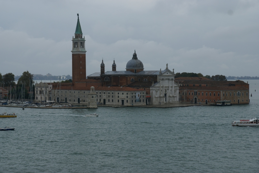 Looking from an elevated floor at a large structure with a white front and red brick church and attached dormitories for an abbey fills an entire island in the Venice lagoon