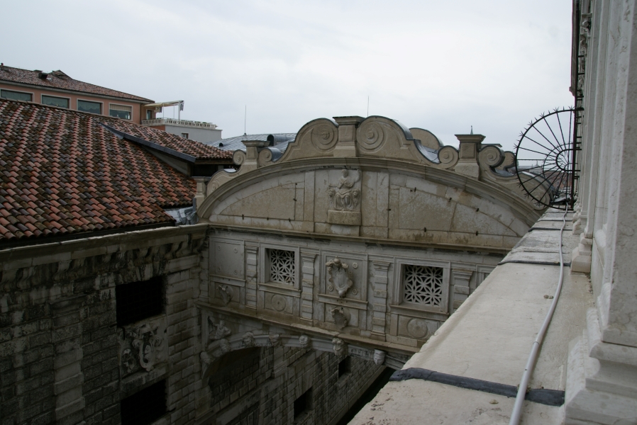 Leaning out a window on an upper floor looking at an enclosed stone bridge between the upper floors of two buildings with small windows and carved flower shaped grills