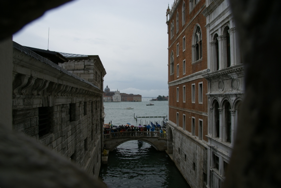 Looking out of a small opening in carved stone as a canal passes between two buildings then under a low bridge and passes into a large body of water with boats and developed islands in the distance