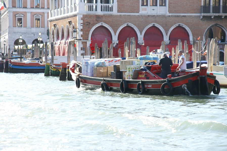 A flat boat filled with boxes of produce and other household goods maneuvers past other boats moored at piles along a canal