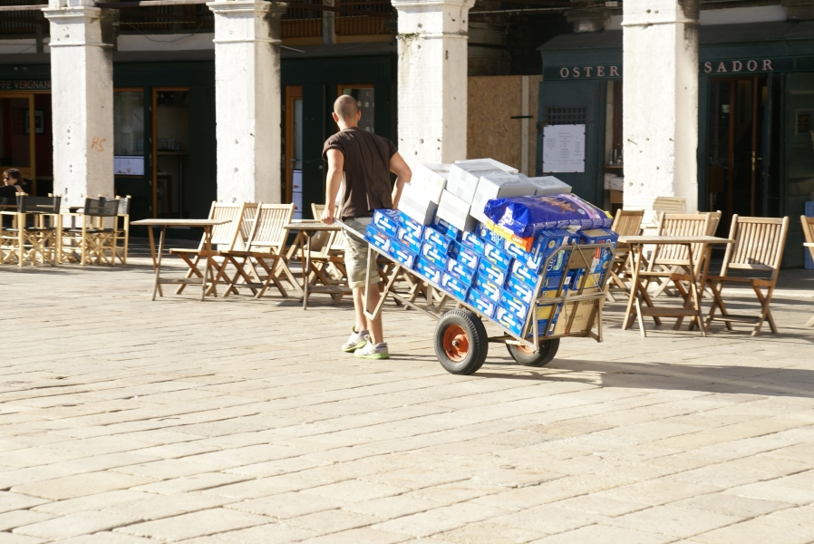 A man walks pulling a two-wheeled cart laden with over 100 pounds worth of boxes of cat food and litter past wooden chairs set out on a stone paver plaza
