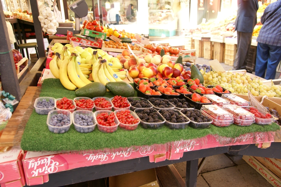 A table in an outdoor market has fresh blueberries, cherries, blackberries, strawberries, bananas, grapes, peaches, pears, and avocados