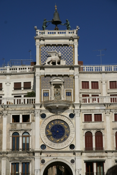 A large bell with two patinaed bronze figures stands atop a tower with a winged lion figure on then different clock mechanisms representing the date, time, and astrological information