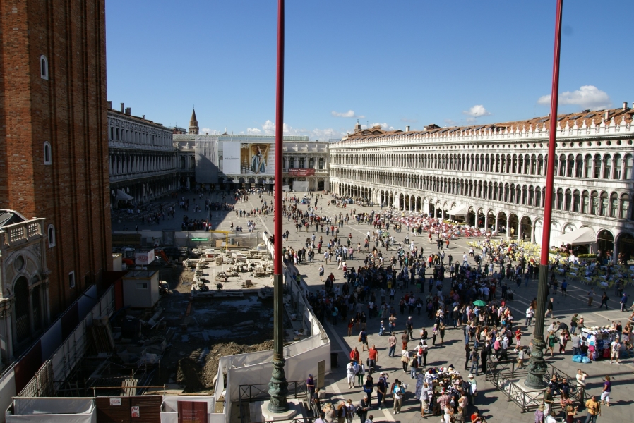 People mill around a very large plaza surrounded on three sides by three-story buildings with arched walls