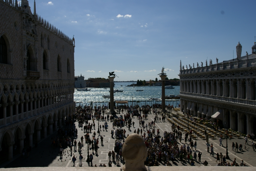 Sun light sparkles off the water as boats pass between islands beyond a large plaza filled with people between buildings and two columns marking the historic entrance to Venice