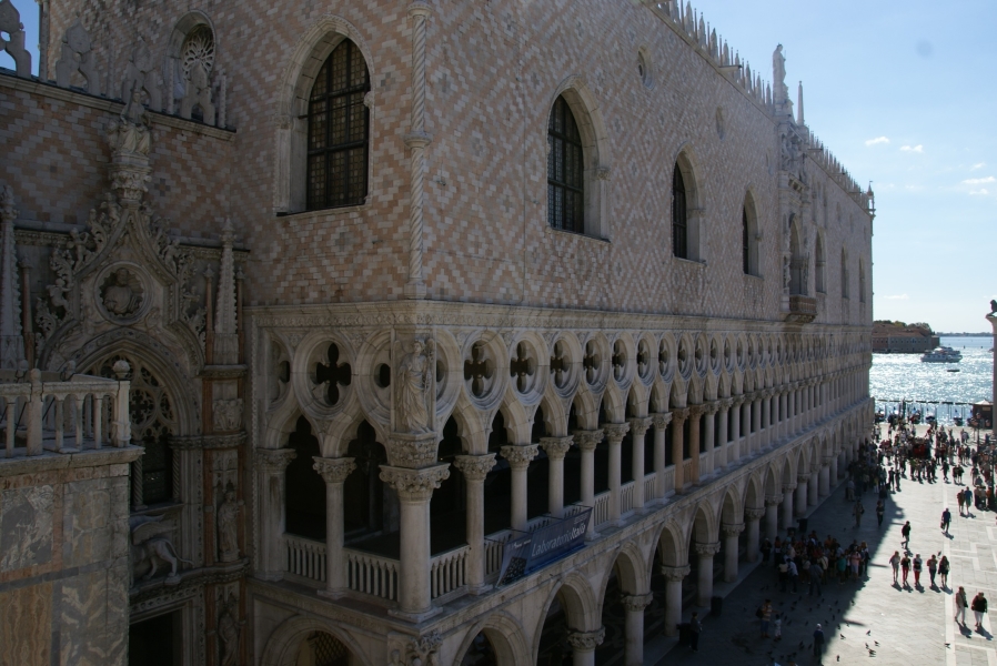 Pink and white stones in a diamond pattern fill the wall above two levels of arched covered walkways of the face of the Doge’s Palace overlooking St. Mark’s Square