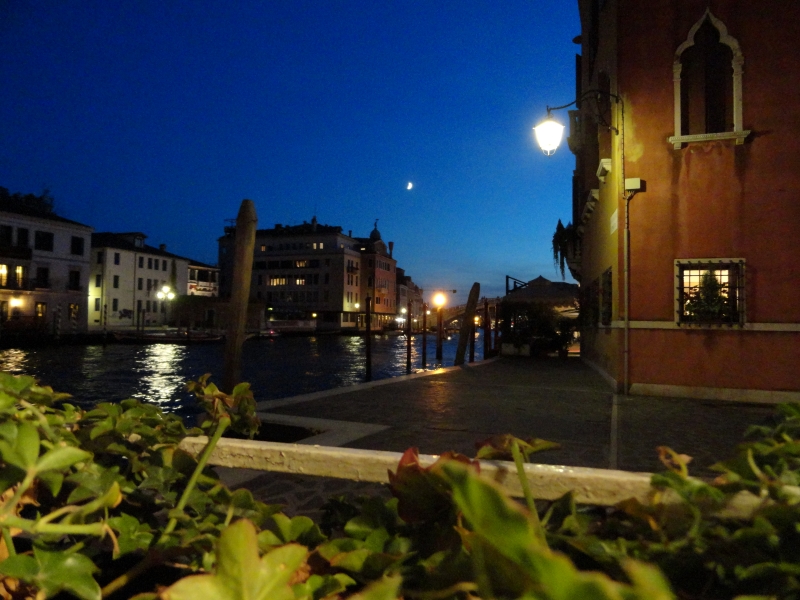 The moon rises in a dark sky with the lights in windows of buildings on either side reflecting on the calm waters of a canal
