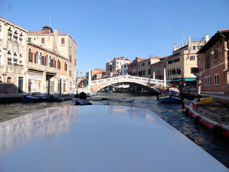 A boat approaches a red stone bridge with bright white railing and columns spanning a canal between three- and four-story buildings