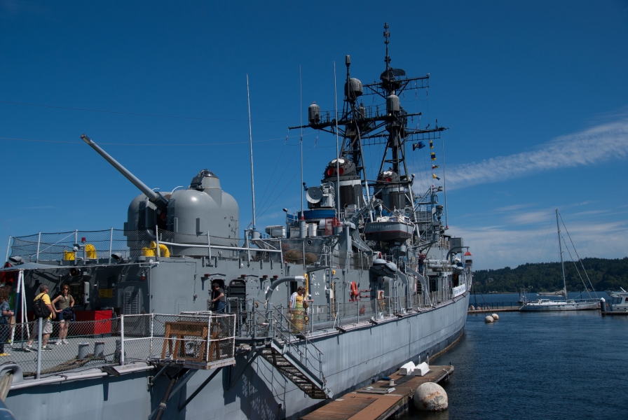 People in casual clothes stand on board a blueish-gray ship bristling with antenna, tall radio masts, and a rotating turret for a gun that fired 8” shells