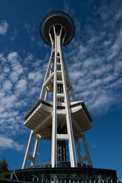 Light fluffy clouds in a blue sky seem to be swirling around a round structure at the top of three sloped trusses and an elevator core running up the middle