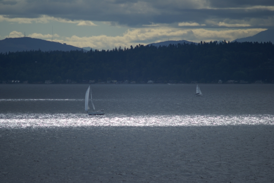 A band of bright, sunlit water passes in front of a sailboat out on the water with hills of green trees rising beyond in the shade of cloudy skies