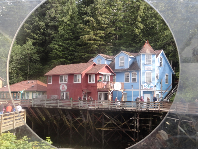 A boardwalk stands on stilts running down into the water in front of a bright blue three-story and a red two-story building with tall thick pine trees beyond