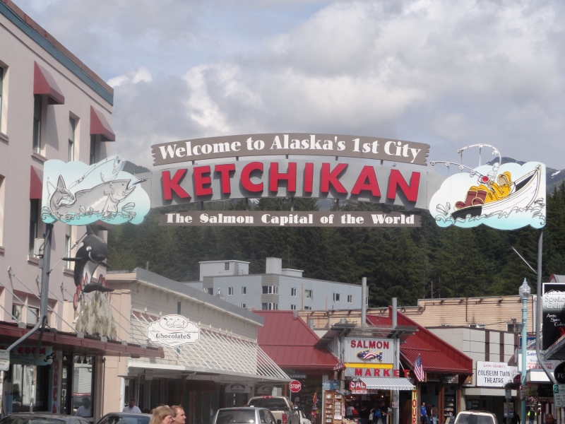 A sign reading “Welcome to Alaska’s 1st City, Ketchikan, The Salmon Capital of the World” crosses the road and shows a fish with a hook in its mouth on one side and a fisherman in a boat on the other