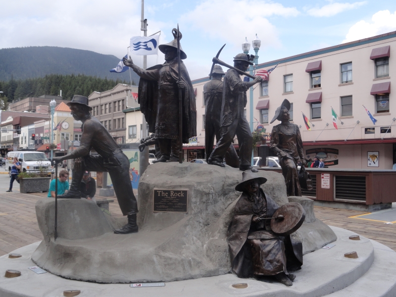 A large concrete stone stands in a plaza with bronze figures wearing the garb and handling tools identify the many people who have contributed to the history of Ketchikan