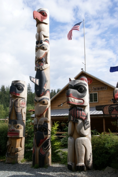 Two short and one tall pole with carvings of different animals stand in front of a wood clad building flying an American flag on a tall flagpole on the roof