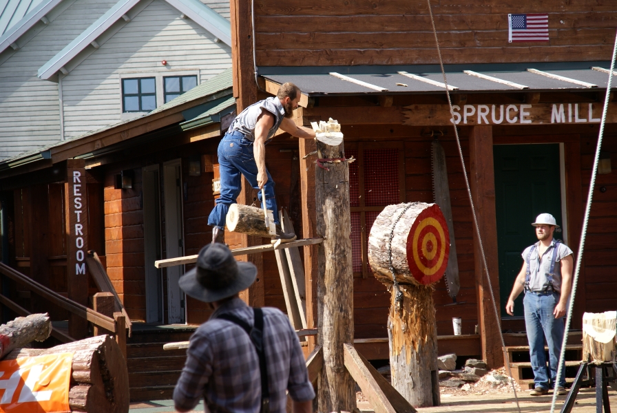 The top of a log held vertically is stuck to the axe head held by a man standing on a wood plank wedged into a lower portion of the same log