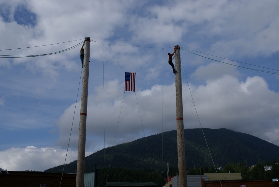 Two tall poles with a climber near the top of each are braced by guide wires stand tall towards the partly cloudy skies with a green, tree covered hill as a backdrop