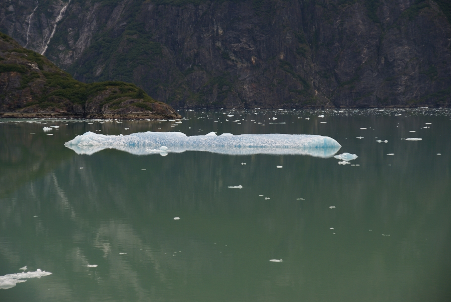 A large chunk of blue ice floats in waters so still the rocky cliffs are evident in the reflection