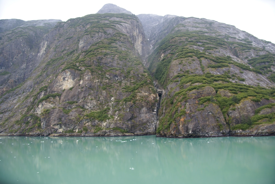 Stone cliffs streaked with moss and lichens drop into smooth green water dotted with small chucks of floating ice 