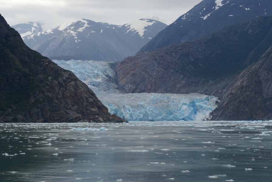 Bluish white ice with a layer of dirt on top rises out of the water and runs back along stone walls with no green towards snow capped mountains in the distance