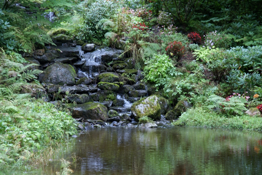 Red and pink flowering plants missed with ferns line the edge of a small, stacked stone waterfall feeding a pond