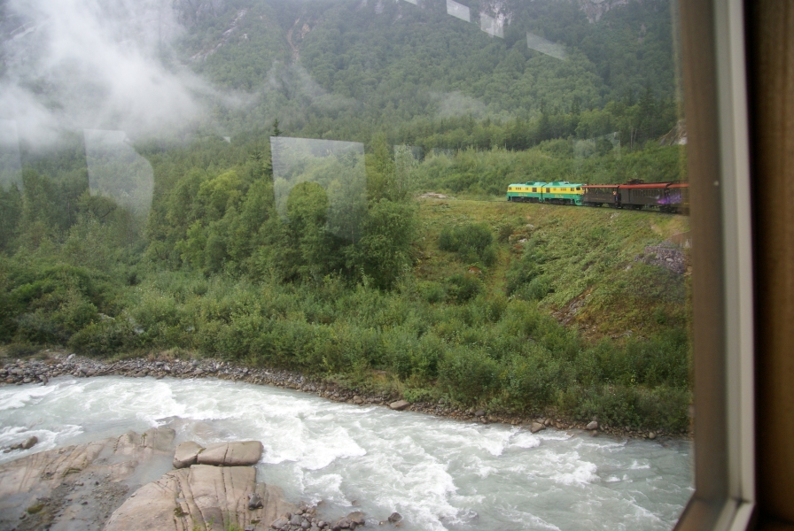 Low clouds sift through pine trees on a hillside as brown train cars with red roofs are pulled by a pair of green and yellow engines across a bridge passing over rushing gray water