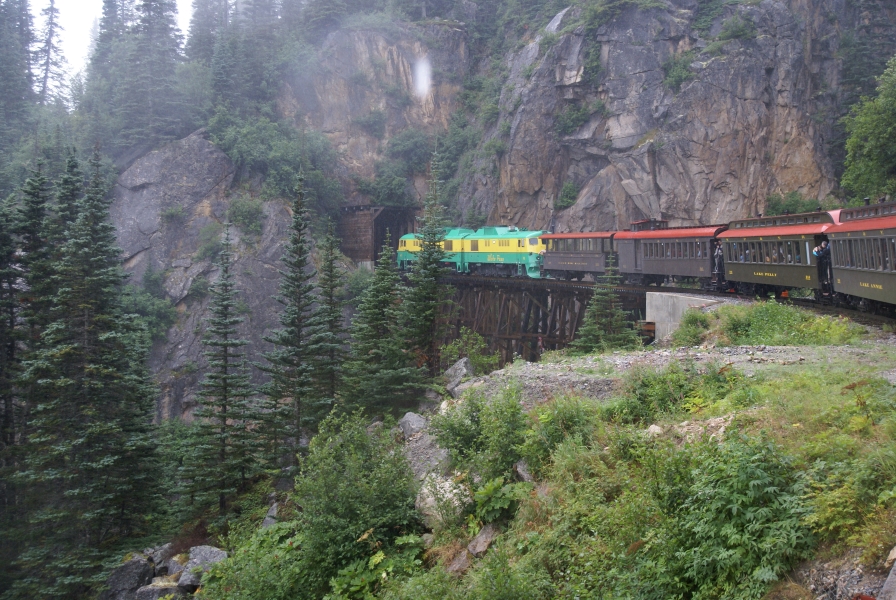 Green and yellow train engines pass over a wood trestle bridge built near a cliff face of broken stone just before passing into a tunnel mouth