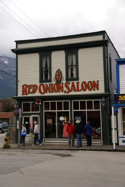 A two-story building with mostly glass on the main floor and yellowish-white board siding and dark green trim sets on a wood board sidewalk with bright red paint identifying it as the “Red Onion Saloon”
