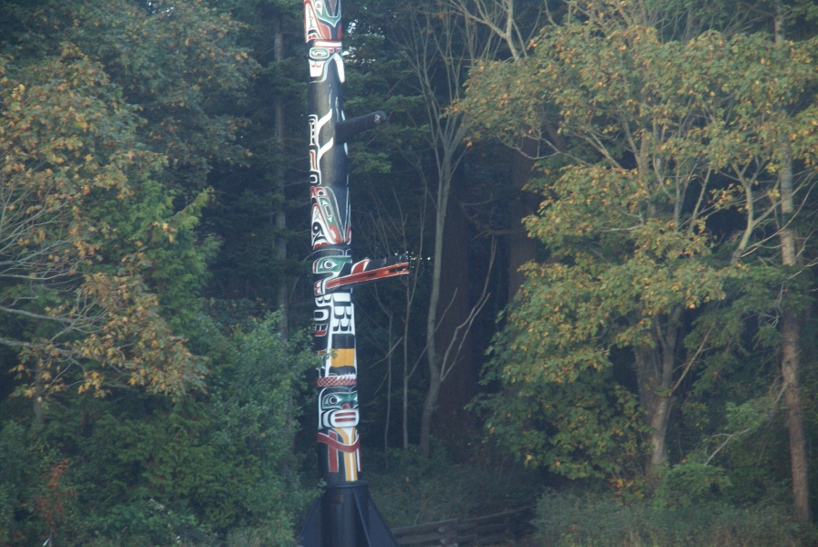 A round pole painted with shades of yellow, red, green and white creating likenesses of different animals sets in a stand of trees