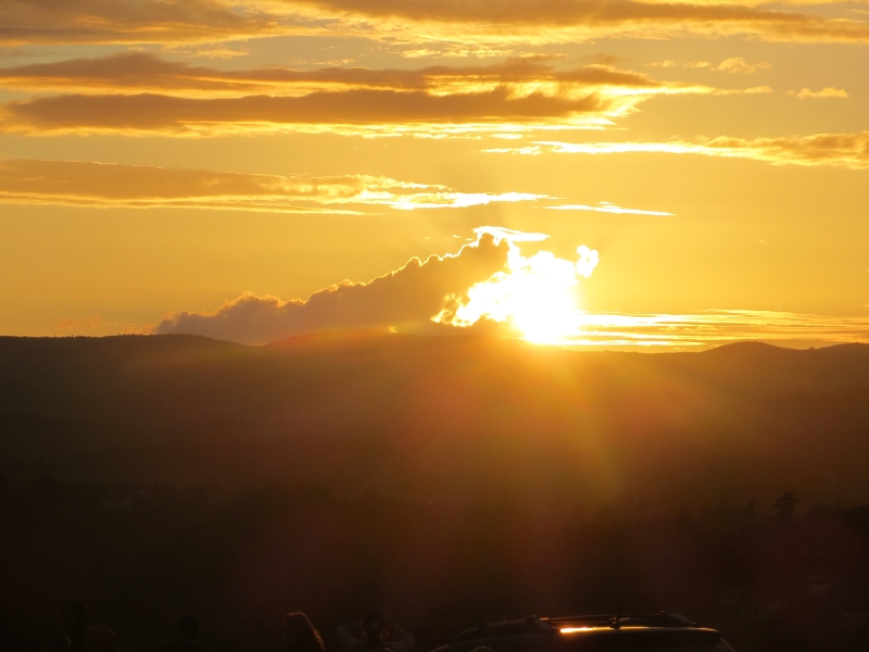 A bright sun sets behind rolling hills bathing the scattered clouds in shades of yellow and gold