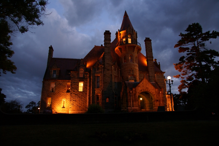 A large stone building with multiple stories, steep roofs, and a round tower near the entry is lit by yellow lights below puffy gray clouds gives off an ominous vibe