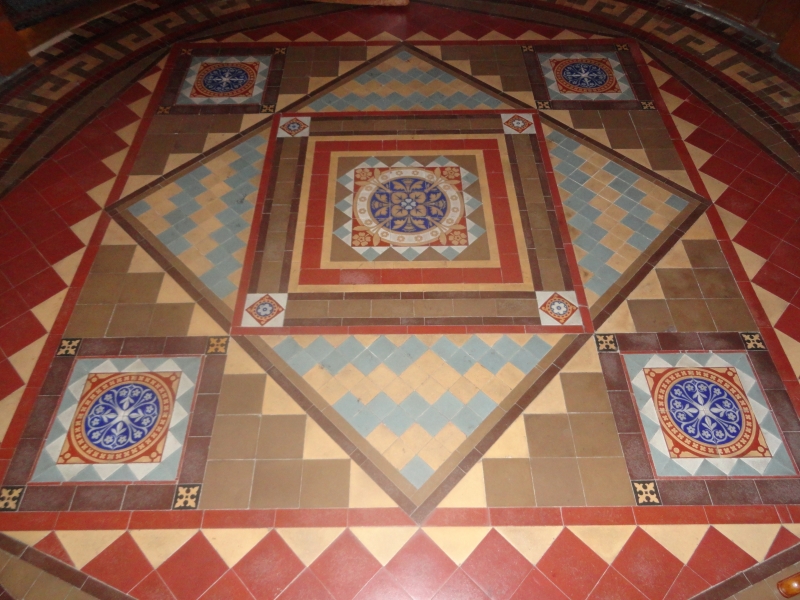 A mosaic on the floor is made from red, brown, light and dark tan, blue, and white tiles accenting patterned tiles of bright blue, white, red and tan flowering motifs