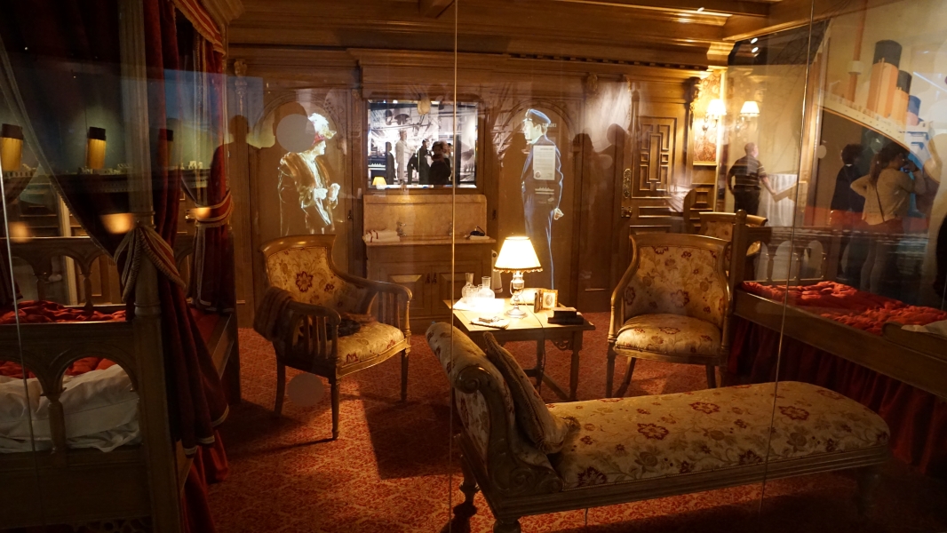 Ornate wood furniture behind glass has two people projected in erly 1900s period clothes