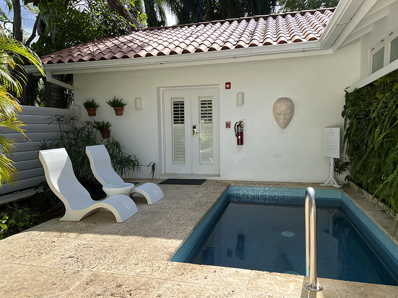 A white building with a small pool in front and a fountain behind two lounge chairs