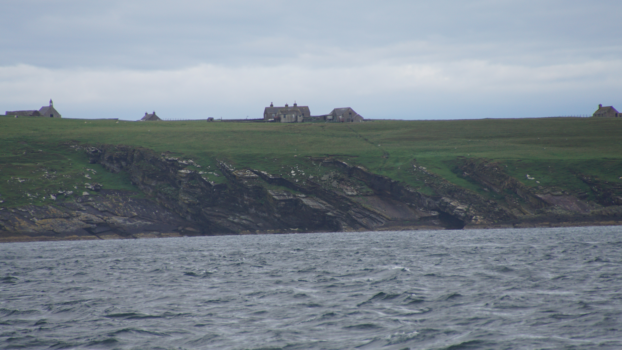 Stone buildings on an open grass plain above a cliff dropping to water