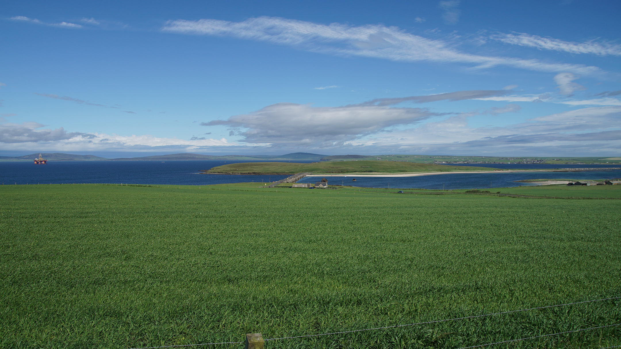 Green grass leading down to a barrier crossing dark blue water with red oil rig in distance
