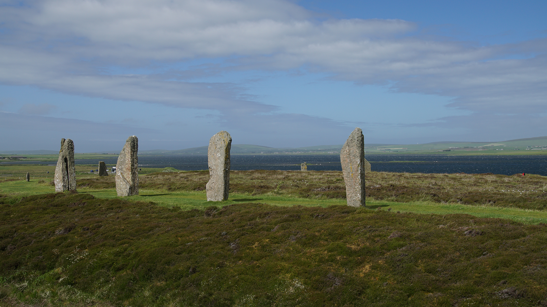 Standing stone markers in a circle on a green field near a lake