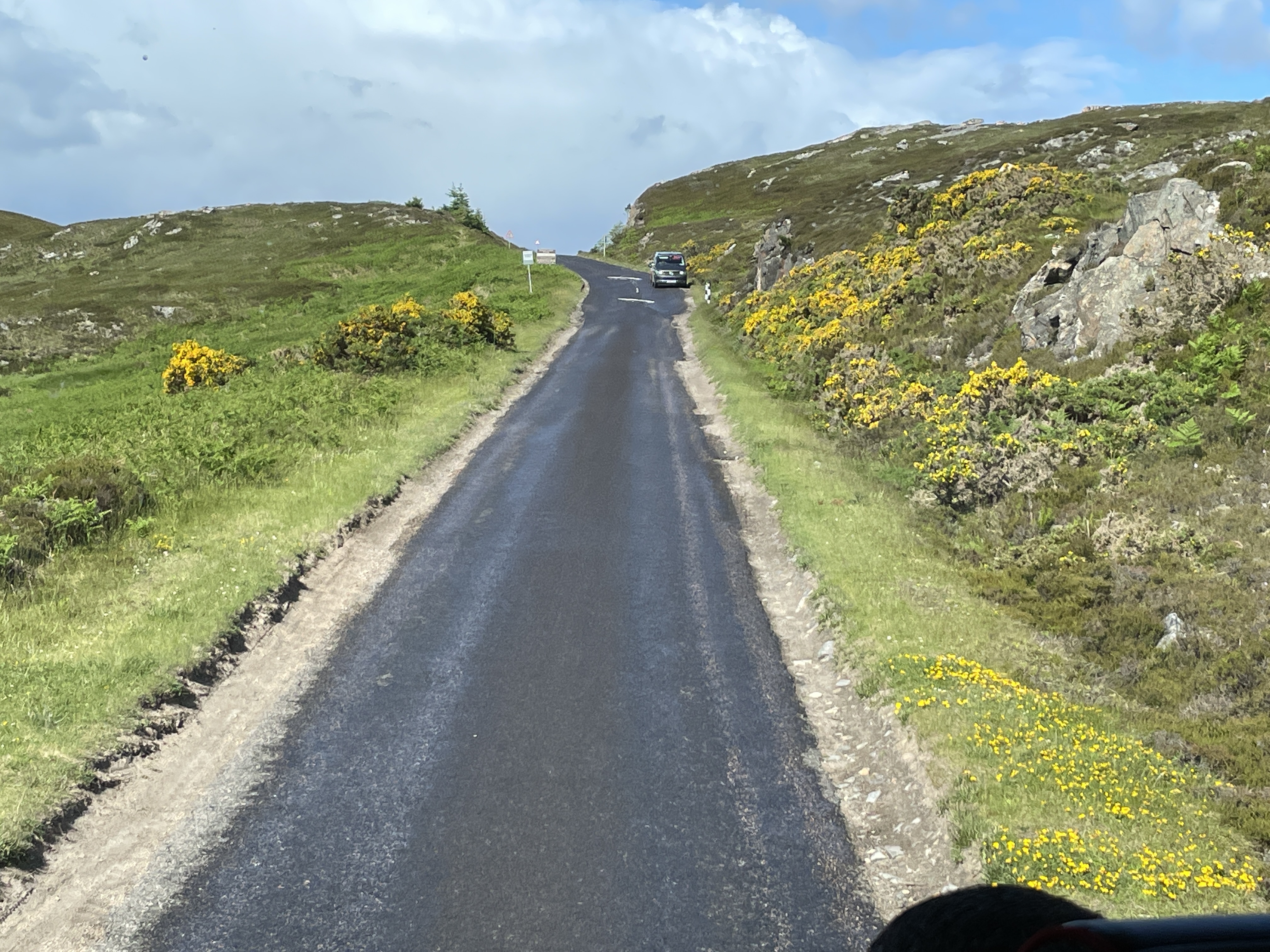 A narrow blacktop road runs through green hills while a car waits for the coach to pass