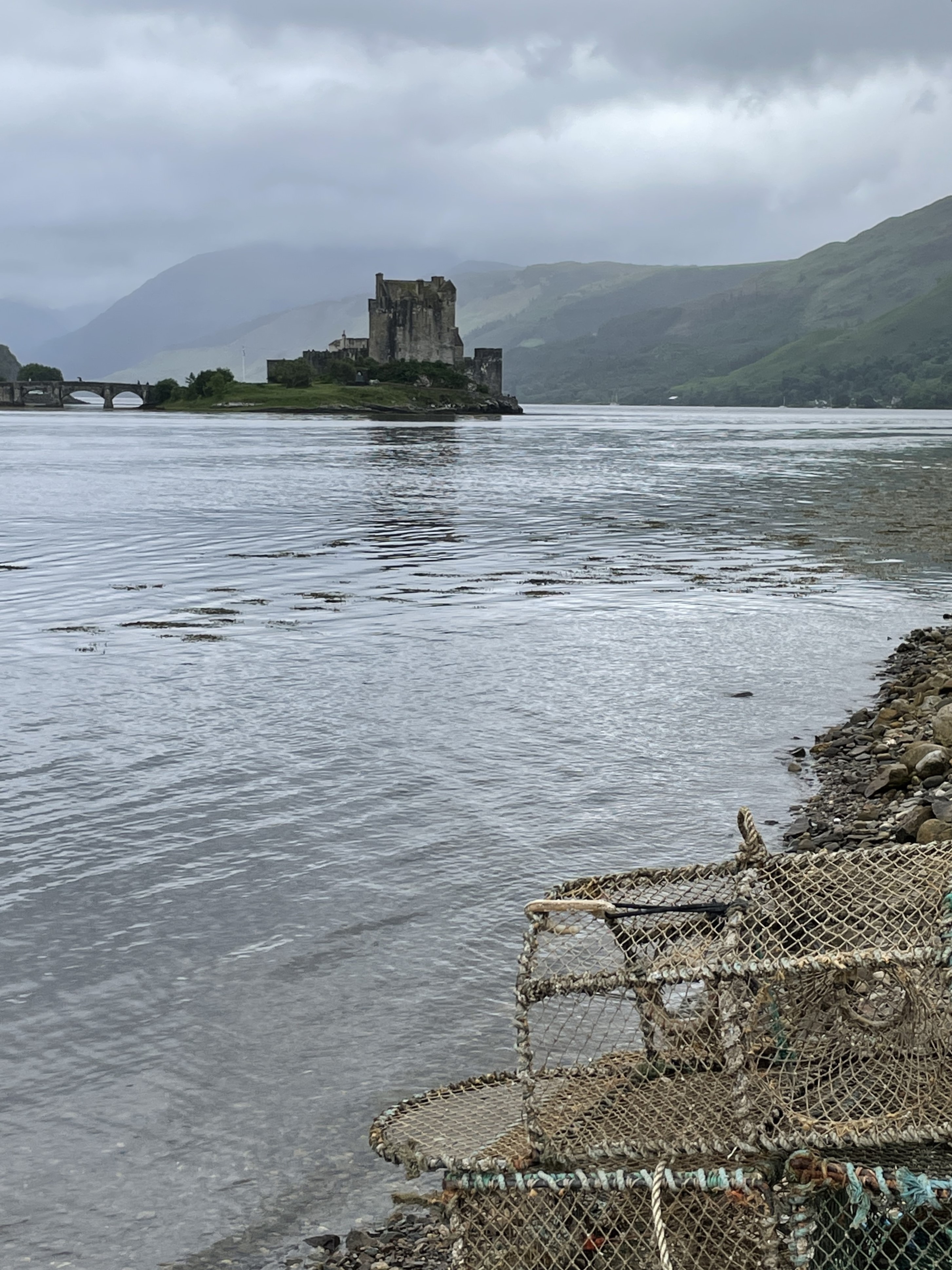 A gray stone castle sits on an island in the middle of a lake surrounded by tall green hills