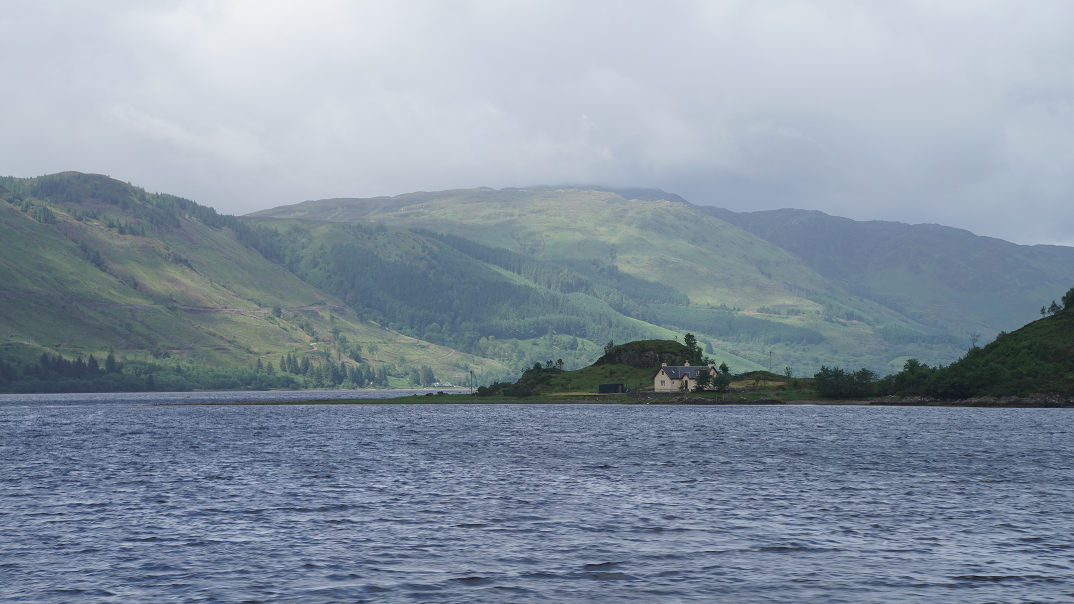 A house sits on the edge of a lake surrounded by green hills with spots of bright sun